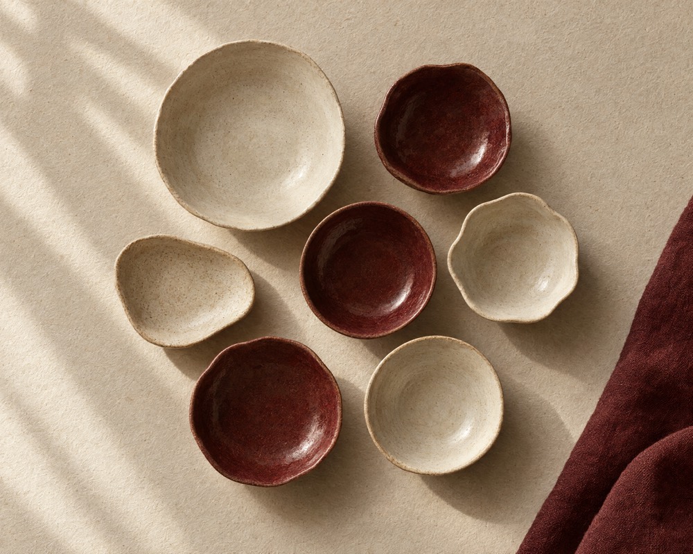 Concept photograph: seven empty handmade ceramic dishes in cream and oxblood glazes arranged together on cream paper